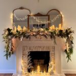 Decorated white fireplace mantel with green garlands, glowing candles, and fairy light mirrors.