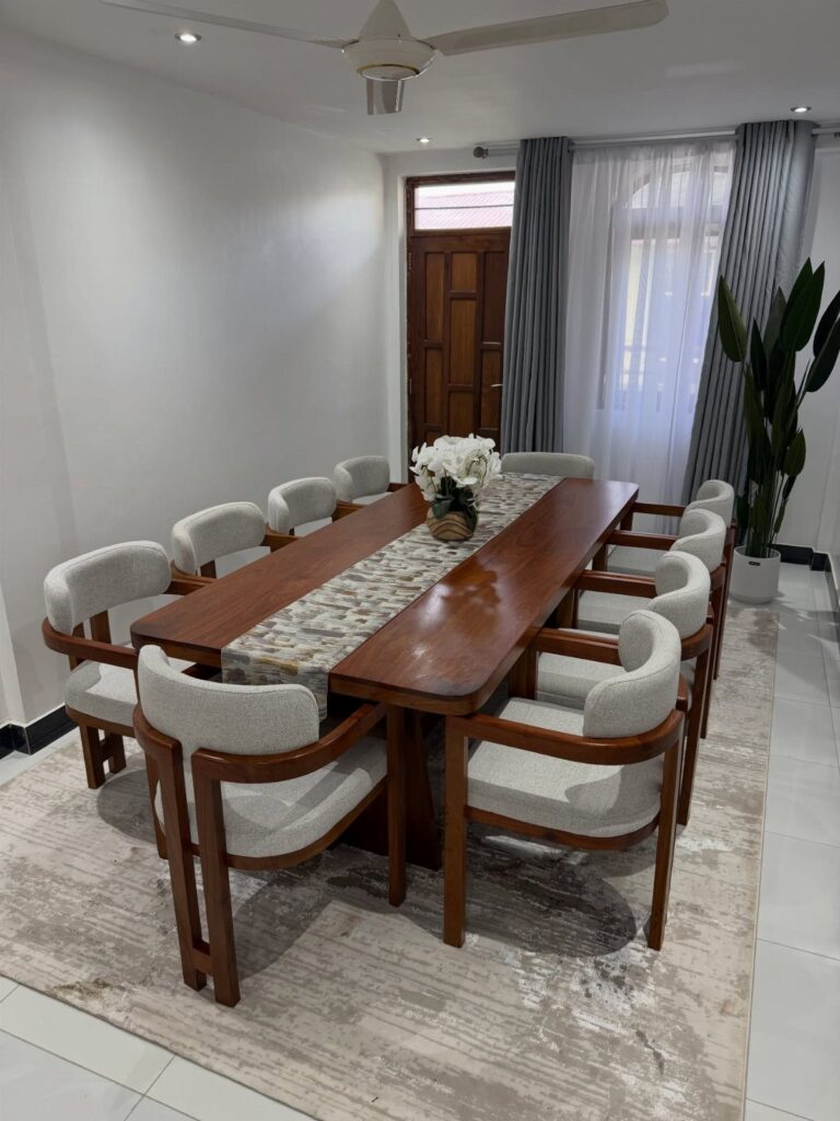 Long wooden dining table with eight grey upholstered chairs and white floral centerpiece.