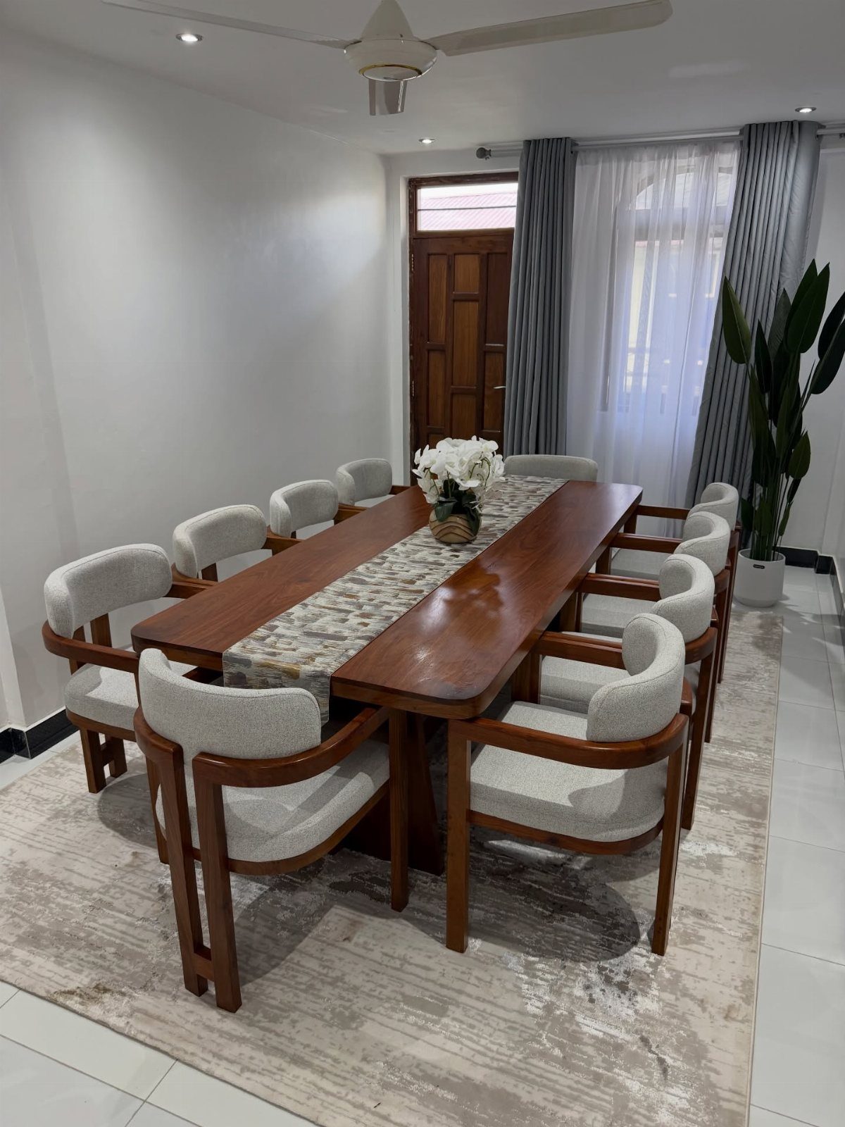 Long wooden dining table with eight grey upholstered chairs and white floral centerpiece.