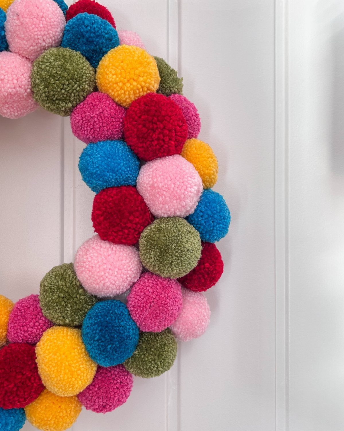 Colorful pom-pom wreath hanging on a white paneled wooden wall.