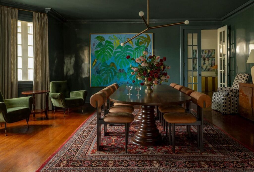 Dining room with dark green walls, oval wood table, and large leaf painting.