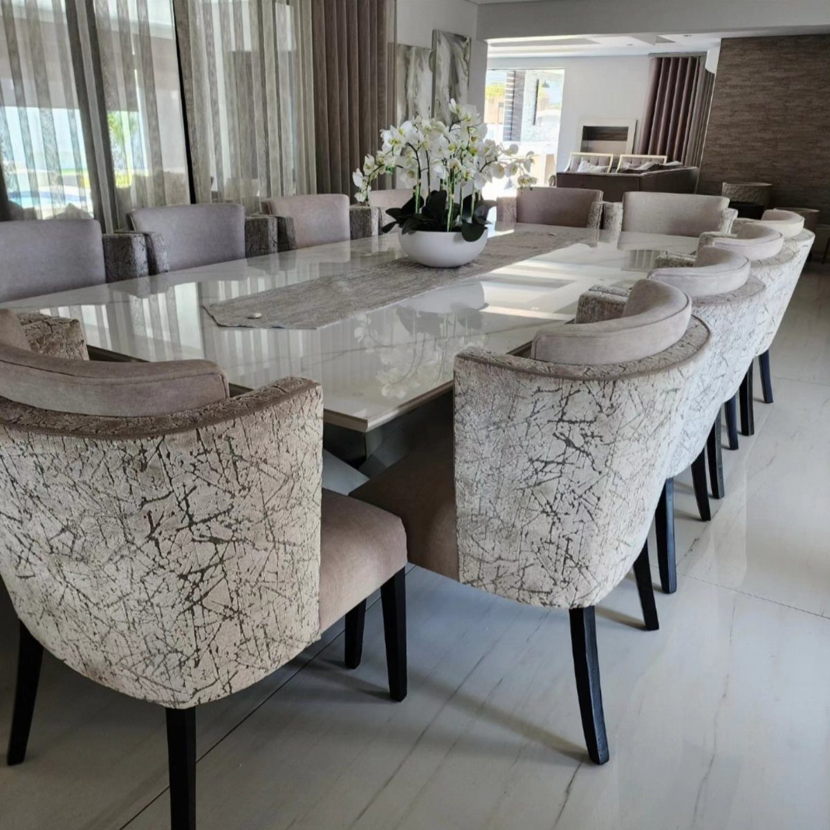 Long white glossy dining table with twelve marbled beige upholstered chairs and white orchids.