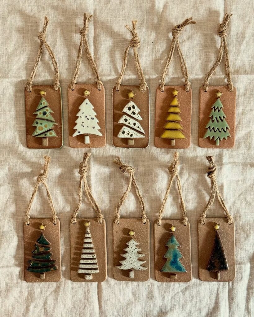 Ten handmade clay Christmas ornaments with colorful tree designs on a white fabric.