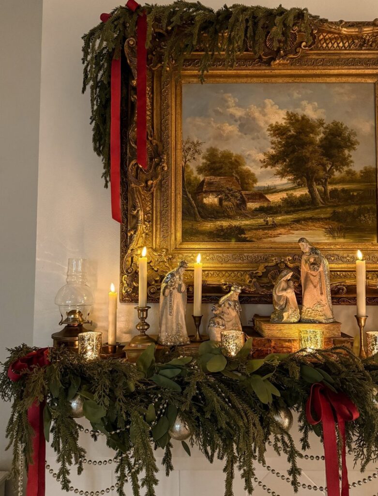 Gold-framed painting decorated with evergreen garlands, red ribbons, and white candles on a mantel.