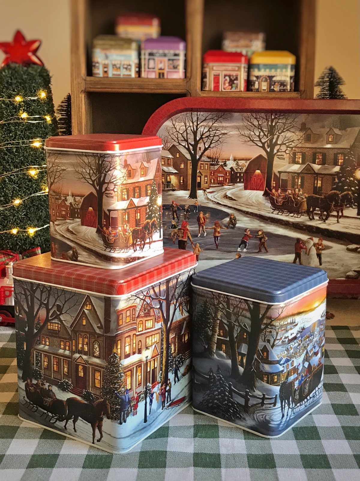 Vintage Christmas tins with winter village scenes arranged on a checkered tablecloth.